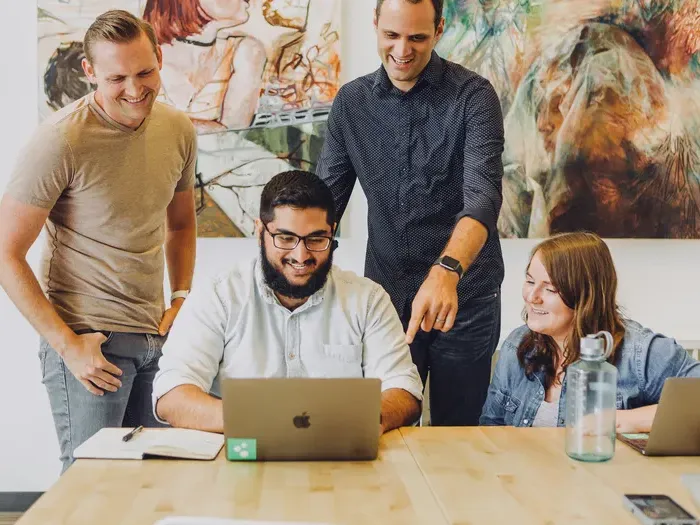 Smiling coworkers gather around a laptop in a bright office, collaborating to review profiles and improve listings